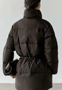 Czarna pikowana kurtka puchowa z wysokim kołnierzem i taliowaniem w pasie. Posiada przeszywane sekcje i gładką teksturę. Modelka trzyma czarną kopertówkę.