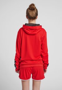 Rode sportieve hoodie van textuurstof, met een zwart omrand capuchon en bijpassende rode shorts met witte rand. Achteraanzicht toont ontwerpinformatie.
