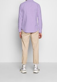 Camicia a maniche lunghe lavanda con orlo curvo, abbinata a chinos beige arrotolati e sneaker grigie. Tessuto uniforme, senza motivi visibili.