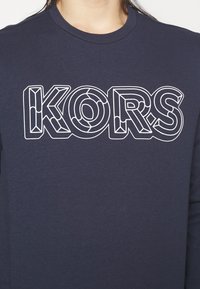 Námorně modré bavlněné tričko s velkým, bílým obrysem loga "KORS" s geometrickými detaily. Kulatý výstřih a dlouhé rukávy.