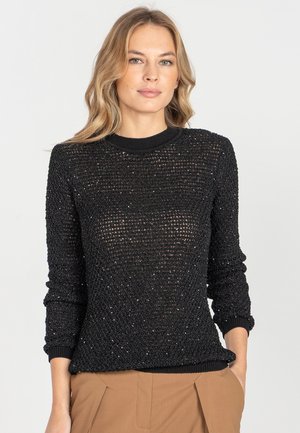 Kobieta z blond włosami, ubrana w czarny sweter o fakturze z długim rękawem i jasnobrązowe spodnie, stoi na tle białego tła.