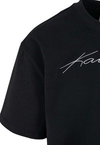 T-shirt noir à manches courtes. Présente un texte brodé blanc "Kanu" sur le devant. Fabriqué en coton doux avec un col rond.