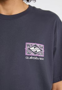 Tamsiai mėlynos spalvos medvilninis marškinėlis su baltu ir rožiniu Quiksilver logotipu, įkomponuotu į raštuotą rombo formą. Trumpomis rankovėmis ir apvaliu kaklu.