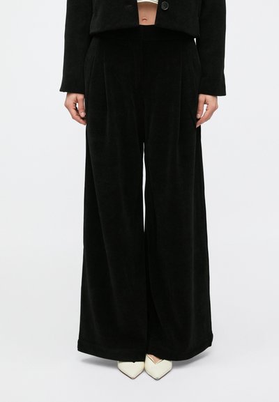 Personne portant un pantalon noir taille haute à jambes larges, une veste noire courte et des chaussures blanches à bout pointu sur un fond uni.
