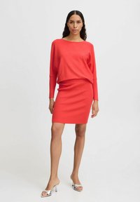 Pull en tricot rouge à coupe ample associé à une jupe à côtes assortie. Le modèle porte des sandales à talons argentées. Design simple et sans fioritures.