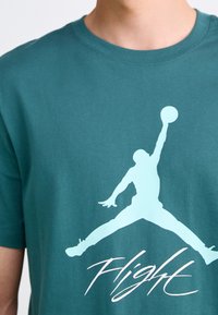 Osoba oblečená v tyrkysovém tričku s siluetou basketbalisty světle modré barvy, který drží míč, a pod ním nápis "Flight" v kurzívě.