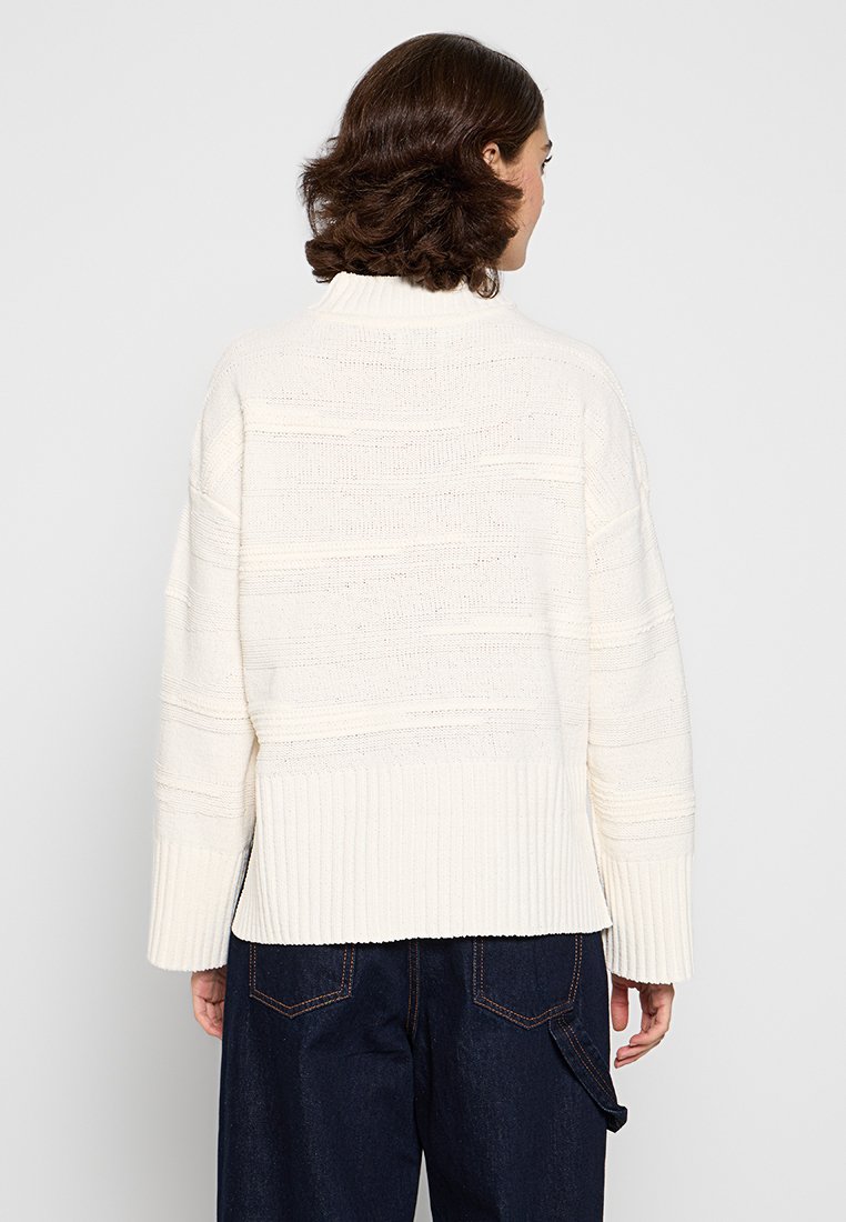Suéter crema con un diseño texturizado y acanalado y mangas anchas. Presenta rayas horizontales en la parte posterior y un cuello alto.