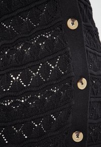 Îmbrăcăminte tricotată neagră, cu un model texturat, având mari butoane decorative pe o latură. Materialul are un design stratificat, cu tricot deschis.