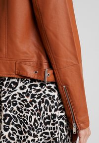 Giacca in pelle marrone con vestibilità slim. Presenta una zip laterale, chiusura a scatto e una texture liscia. Abbinata a una gonna a stampa leopardo.
