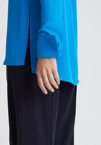 Langärmliges, leuchtend blaues Hemd mit glatter Textur. Verfügt über geknöpfte Manschetten und Seitenschlitze. Kombiniert mit dunklen, maßgeschneiderten Hosen.