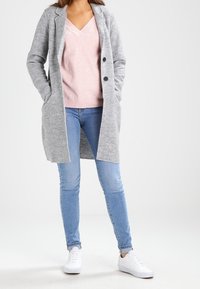 Abrigo largo gris con cuello, cierre de botones, usado sobre un suéter de pico rosa claro. Jeans ajustados de azul claro y zapatillas blancas completan el conjunto.