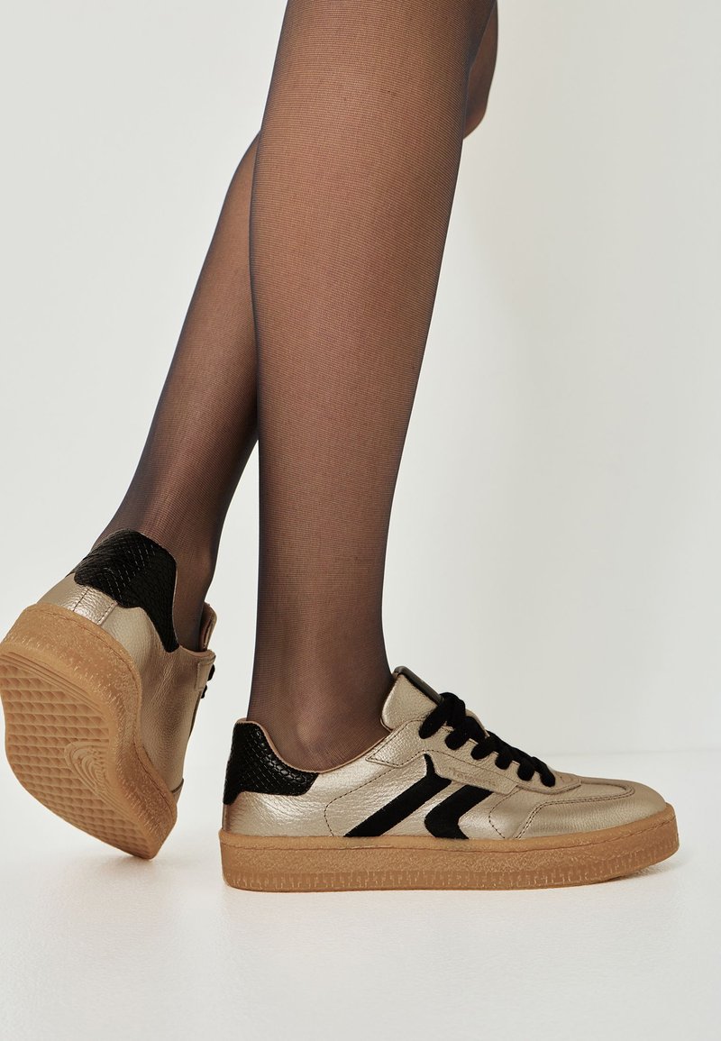 Metaliczne złote sneakersy z czarnymi akcentami, klasyczny sznurowany design, teksturowany detal z imitacji wężowej skóry na pięcie oraz gumowa podeszwa.
