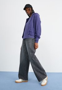 Corta-vento roxo com fecho éclair, de corte oversized, combinado com jeans pretos de cintura alta e ténis brancos com solas bege. Boné preto usado para trás.