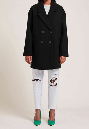 Osoba ubrana w czarny dwurzędowy płaszcz, jasnoniebieskie podarte dżinsy oraz jaskrawozielone szpilki, stojąca na jednolitym tle.