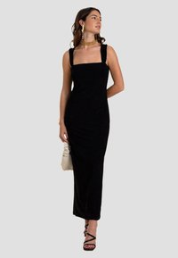 Zwarte mouwloze maxi-jurk met een vierkant decolleté, zacht materiaal, strakke pasvorm en minimale details. Combinatie met zwarte sandaaltjes met bandjes.