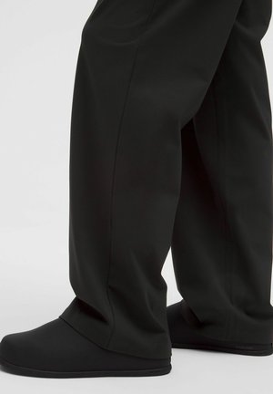 Osoba u crnim širokim hlačama i crnim cipelama bez vezivanja stoji na svijetloj površini.