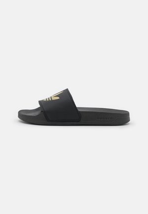 Tongs noires Adidas avec semelle texturée et logo trefoil doré sur la large sangle, montrées en vue latérale sur fond blanc.