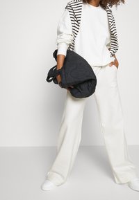 Witte oversized sweatshirt, crèmekleurige broek met wijde pijpen, zwarte gewatteerde tas en witte sneakers. Gestreepte hoodie nonchalant over de schouder gedrapeerd.