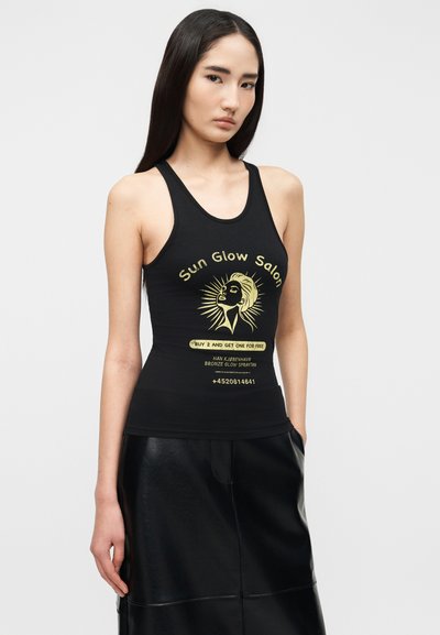 Γυναίκα με μακριά σκουρόχρωμα μαλλιά, που φοράει μαύρο αμάνικο τοπ με κίτρινο γραφικό "Sun Glow Salon" και μαύρη δερμάτινη φούστα, στέκεται μπροστά σε απλό φόντο.