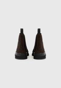 Botas de ante marrón hasta el tobillo con paneles elásticos laterales, suelas de goma negras y tiradores para facilitar el uso. La superficie texturizada añade interés visual.