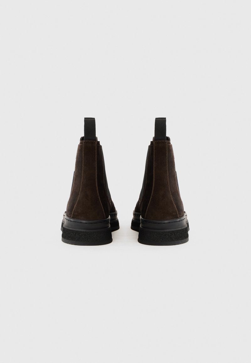 Stivali alla caviglia in suede marrone con pannelli elastici a lato, suole in gomma nera e linguette per una facile vestibilità. La superficie testurizzata aggiunge interesse visivo.