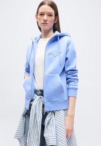 Νεαρή γυναίκα που φοράει ένα ανοιχτό γαλάζιο φούτερ με φερμουάρ πάνω από ένα λευκό πουκάμισο, με μια ριγέ μπλούζα δεμένη γύρω από τη μέση της και μπλε τζιν.