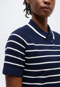 Άτομο που φοράει μπλε ναυτικό πλεκτό πόλο πουκάμισο με λευκές οριζόντιες ρίγες και τρία κουμπιά, κοντά μανίκια και γιακά με λευκή επένδυση.