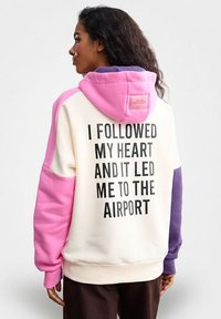 Bluza z kapturem w modnym stylu color-blocking, z białym korpusem oraz różowymi, fioletowymi i beżowymi akcentami. Na plecach napis: "PODSŁUCHAŁEM SWOJE SERCE I ZAPROWADZIŁO MNIE NA LOTNISKO."