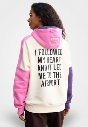 Bluza z kapturem w modnym stylu color-blocking, z białym korpusem oraz różowymi, fioletowymi i beżowymi akcentami. Na plecach napis: "PODSŁUCHAŁEM SWOJE SERCE I ZAPROWADZIŁO MNIE NA LOTNISKO."