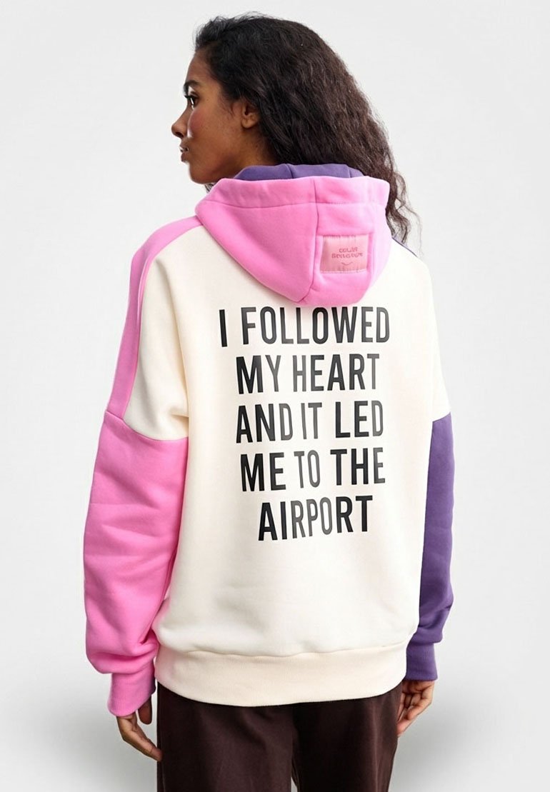Bluza z kapturem w modnym stylu color-blocking, z białym korpusem oraz różowymi, fioletowymi i beżowymi akcentami. Na plecach napis: "PODSŁUCHAŁEM SWOJE SERCE I ZAPROWADZIŁO MNIE NA LOTNISKO."