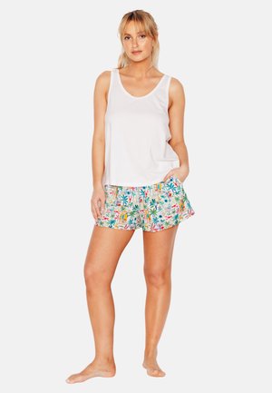 Donna in piedi a piedi nudi che indossa una canottiera bianca e pantaloncini colorati con stampa tropicale, una mano in tasca, posa rilassata.