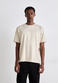 Beige bomull t-skjorte med korte ermer, rund hals og preget "BOSS"-logo på brystet. Kombinert med svarte bukser.