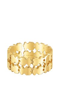 Bransoletka w kolorze złotym z ażurowym wzorem falistych kształtów, gładką teksturą i polerowanym wykończeniem. Wewnątrz wygrawerowano napis 'TOUS S925'.