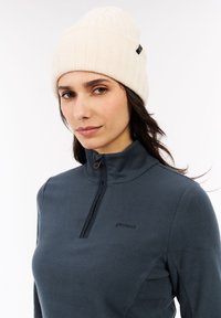 Donna che indossa un berretto di lana color crema e un top in pile grigio scuro con zip parziale, in piedi contro uno sfondo bianco semplice.
