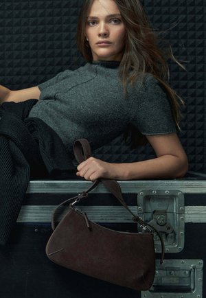 Brązowa zamszowa torebka o zaokrąglonym kształcie, wyposażona w regulowany pasek i detal z zamkiem błyskawicznym. Na tle o fakturze czarnego koloru.