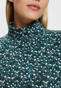 Maglione a collo alto color teal con un motivo a macchie in bianco, nero e toni di grigio. Il tessuto ha una superficie liscia con una media elasticità.