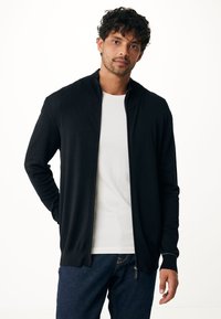 Svart dragkedjetröja med hög krage, tillverkad av ett slätt tyg. Har långa ärmar och en figursydd design, bärs över en vit skjorta.