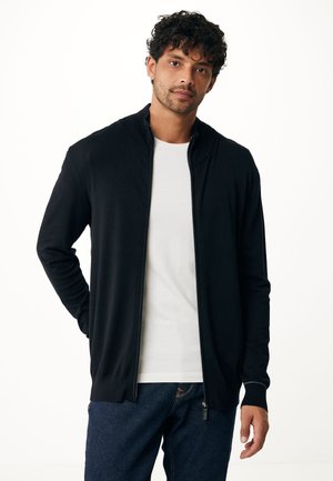 Black zip-up cardigan with a high collar, made of a smooth fabric. Features long sleeves and a fitted design, worn over a white shirt.
