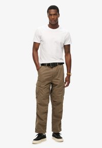 Khaki cargobroek met meerdere zakken, gecombineerd met een eenvoudig wit T-shirt. Model draagt een zwarte riem en zwarte sneakers met een crèmekleurige zool.