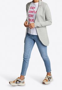 Lichtgrijze blazer over een witte t-shirt met roze tekst, gecombineerd met lichtblauwe jeans en multicolor sneakers met oranje en marineblauwe accenten.
