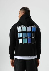 Zwarte hoodie met een raster van twaalf gekleurde vierkanten op de achterkant, gelabeld met Pantone-namen, die verschillende tinten blauw tonen.