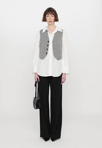 Gilet grigio sopra una camicia bianca con bottoni, abbinato a pantaloni larghi neri. L'outfit include una piccola borsa nera.