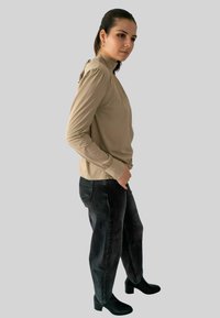 Vrouw in een beige blouse met lange mouwen en hoge nek, met losse zwarte jeans, staande in profiel met de handen in de zakken tegen een effen achtergrond.