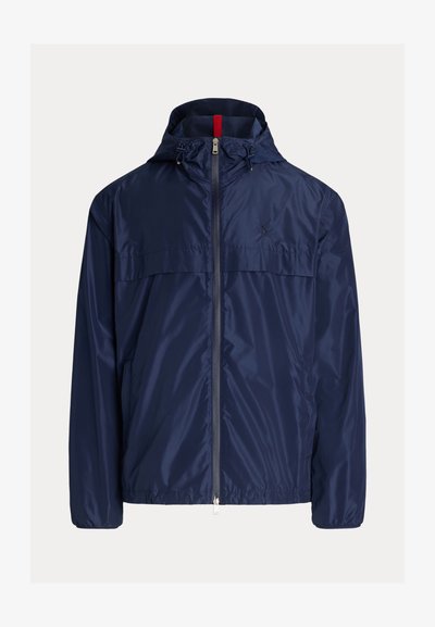 Marineblå vanntett jakke med hette, frontglidelås og elastiske mansjetter. Har et rødt pryd på kragen og lysreflekterende materiale.