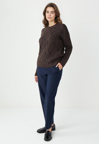 Brązowy sweter o fakturze z wzorem w diamenty, w połączeniu z granatowymi spodniami w talii i czarnymi loafersami, na białym tle.