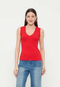 Νεαρή γυναίκα με μακριά σκούρα μαλλιά που φορά χρυσούς κρίκους στα αυτιά, αμάνικο κόκκινο τοπ με V λαιμόκοψη και μπλε τζιν, στέκεται μπροστά σε απλό λευκό φόντο.