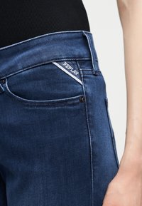 Κοντινό πλάνο της μέσης με σκουρόχρωμο μπλε τζιν Replay και μαύρο τοπ, εστιασμένο στο μικρό λογότυπο Replay κοντά στην μπροστινή τσέπη.