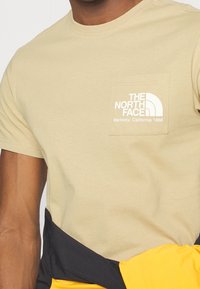 Muž v béžovém tričku The North Face s logem a textem na kapse, držící černou a žlutou bundu kolem pasu.