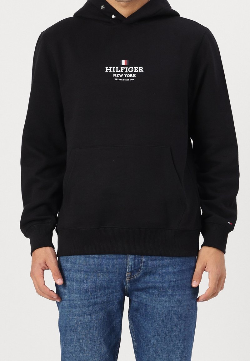 Sweat à capuche noir avec poche avant et logo "Hilfiger New York Established 1985", porté avec un jean bleu sur une personne debout.