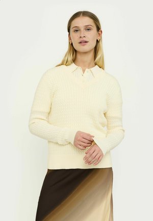 Maglione color crema con scollo a V, caratterizzato da orlo e polsini a coste. Indossato sopra una gonna sfumata beige e marrone.